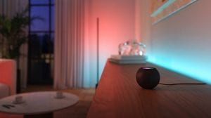 A black smart speaker placed on a wooden surface in front of colored smart lights casting blue and red light on the wall.
