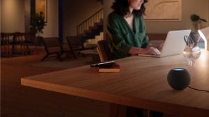 Person working on a laptop at a wooden table in a softly lit home office, with a smart speaker and desk lamp nearby.