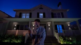 Person standing outside a house at night using a smartphone, with outdoor smart lighting illuminating the porch and garden.
