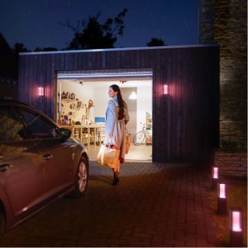 A person carrying shopping bags walks from a car toward a garage at night, illuminated by smart outdoor lighting fixtures.