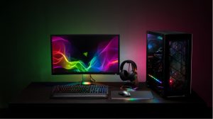 A computer monitor, keyboard, mouse, and headphones on a desk, with RGB lighting effects reflecting on the wall and surfaces.