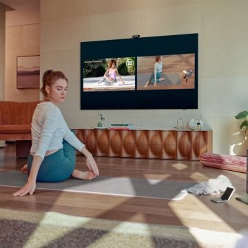 A person practicing yoga in a living room, following an exercise video on a wall-mounted TV with natural light illuminating the space.
