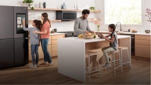 A family in a modern kitchen prepares food, uses a tablet, and interacts with a smart refrigerator under bright ceiling lighting.
