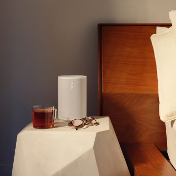 A white Sonos speaker placed on a bedside table next to a glass cup with tea and a pair of eyeglasses, near a wooden headboard.