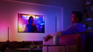 Person watching TV in a living room with smart lighting casting colorful hues on walls and furniture, voice assistant device visible.