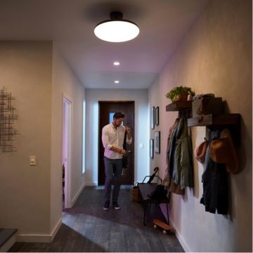 Person entering a hallway, adjusting a bag, with overhead lighting illuminating coats, hats, and a skateboard on the side.