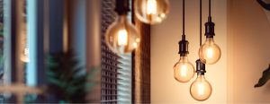 Collection of suspended smart filament bulbs with black sockets, casting light onto nearby wall and window blinds in a living space.