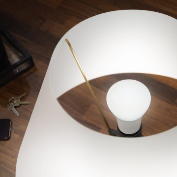 A white smart bulb is installed in a modern lamp with a curved white shade, placed on a wooden floor near keys and a phone.