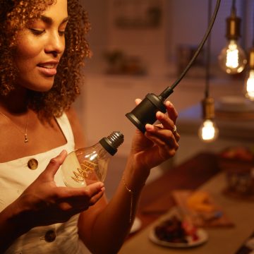 A person installs a filament bulb into a pendant socket in a warmly lit dining room with food on the table in the background.