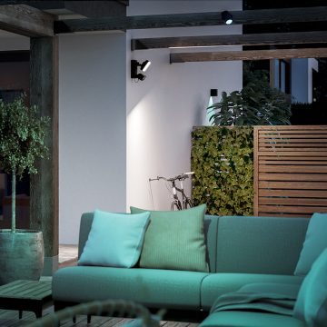 A black outdoor smart floodlight is mounted on a wall, illuminating a patio area with a sofa and visible plants.