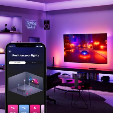 A smartphone displays a lighting control app in front of a TV with colored light syncing, shelves, and a ""Gaming Zone"" sign on the wall.
