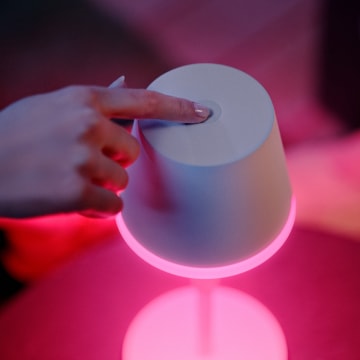 Finger pressing the button on a portable smart lighting device with a glowing pink light ring at the base.