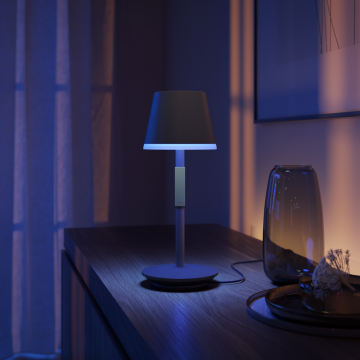 Portable table lamp with a conical shade placed on a wooden surface, emitting blue light onto nearby objects.
