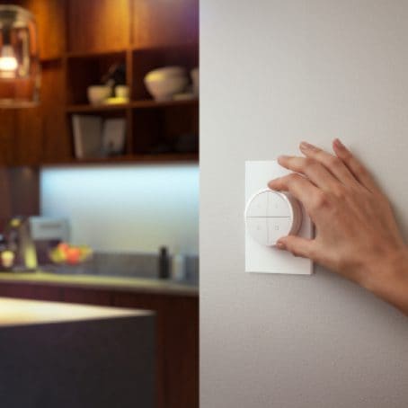 Hand rotating a white smart lighting control dial mounted on a wall, likely to adjust light settings in a kitchen area.