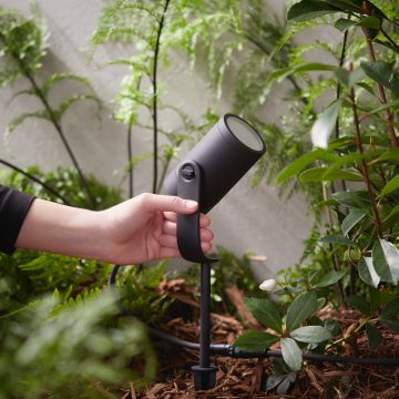 Hand adjusting a black outdoor spotlight in a garden bed among plants, spotlight designed for smart home and smart lights integration.