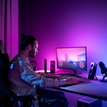 Person using a computer at a desk in a gaming setup, with purple and blue ambient lighting illuminating the workspace.