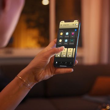 Person using a smartphone app to adjust living room lighting settings in a softly lit home environment during the evening.