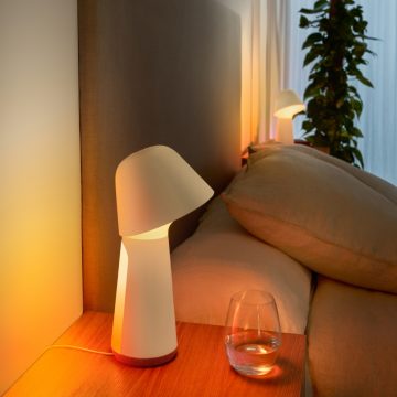 White bedside smart lamp with angled shade on a wooden nightstand, casting light onto the headboard and surrounding area.