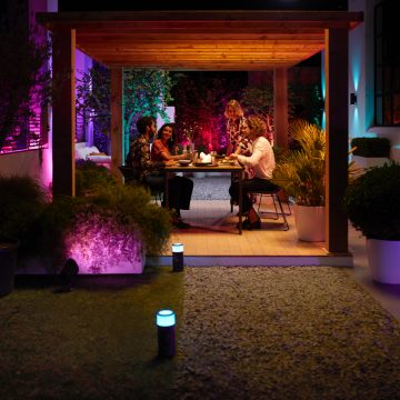 A group of people dining under a wooden pergola on a patio, with colorful outdoor lighting illuminating the garden and seating area.