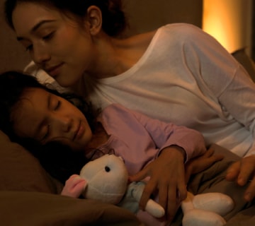 An adult and child rest together on a bed with a plush toy, illuminated by soft, warm ambient lighting in a bedroom setting.