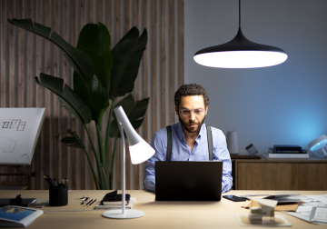 A person working on a laptop at a desk in a modern office, illuminated by indoor LED strip light and desk lamp.
