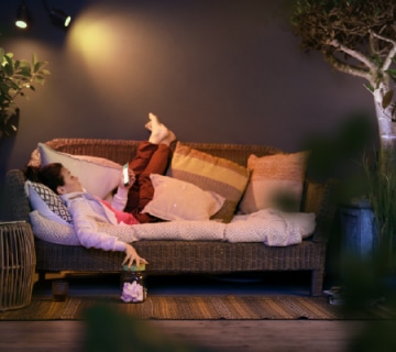 Person relaxing on an outdoor sofa under adjustable smart lighting, holding a phone, surrounded by cushions and plants in a patio setting.