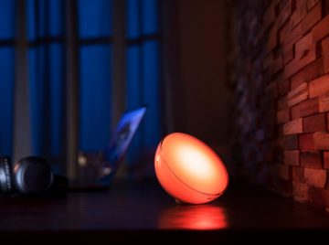 A round smart lamp emitting orange light is placed on a wooden desk near a textured wall, illuminating the surface.