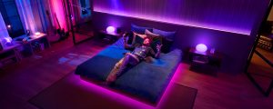 Person relaxing on a bed in a bedroom, illuminated by purple and pink smart lighting, with music equipment nearby.
