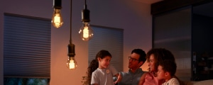 A group of people gather in a living room, sitting together under hanging filament bulbs with warm light.
