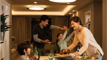 A family gathers around a dining table for a meal in a warmly lit living space with a round LED ceiling light panel overhead.