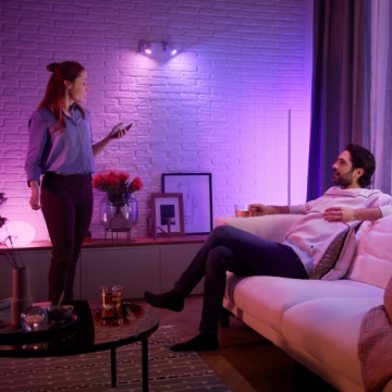 Two people in a living room, one standing with a phone and one seated, with purple smart lighting illuminating the space.