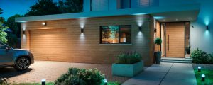 Outdoor security cameras mounted above the garage and entrance, with wall lights illuminating the wooden exterior and driveway.