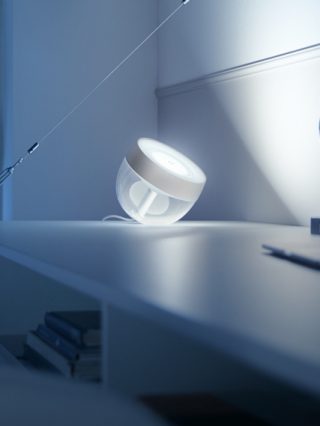 A round smart table lamp with a translucent body placed on a desk, casting white light onto the adjacent wall surface.