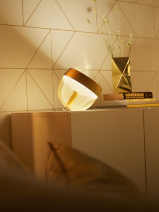 Glass smart table lamp with metallic top placed on cabinet, casting soft white light onto geometric patterned wall and nearby decor.