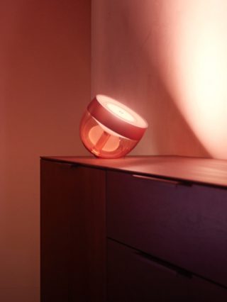 A translucent smart table lamp placed atop a cabinet, casting diffused colored light onto an adjacent wall.