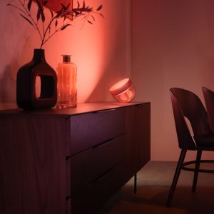 Glass smart table lamp placed on sideboard, casting colored light onto wall behind decorative vases and wooden furniture.