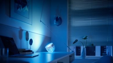 Smart table lamp with translucent body placed on desk, projecting blue light onto nearby wall and workspace in office setting.