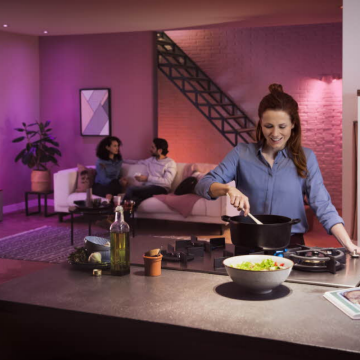 A person cooking at a kitchen island with warm colored smart lighting, while a group sits and talks on a sofa in the background.