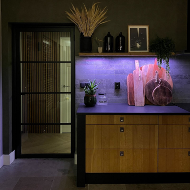 Under-cabinet smart lighting illuminates kitchen counter with cutting boards, plant, and ""HOME MADE"" container against tiled backsplash.