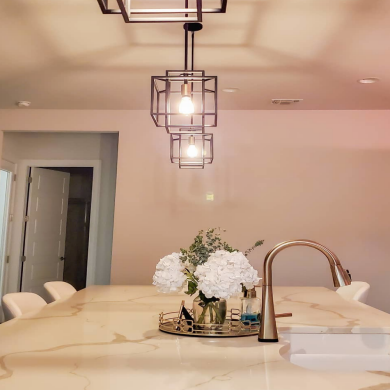Pendant smart bulbs in geometric fixtures hang above a kitchen island, casting light on the marble countertop and sink area.