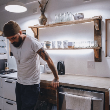 Person standing in a kitchen with under-shelf lighting illuminating dishes and glassware on rustic wooden shelves above a countertop.