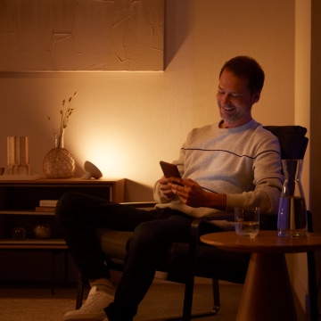 Person seated in a living room using a smartphone, illuminated by soft smart lighting on a sideboard, with water on a nearby table.