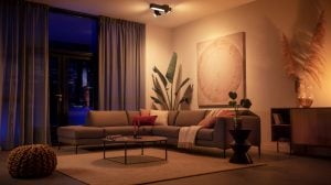 Ceiling-mounted smart lighting fixture illuminating a living room with sectional sofa, wall art, and indoor plants.