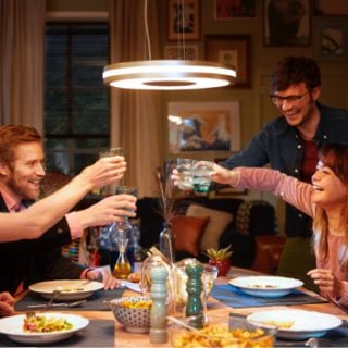 A group shares a meal and raises glasses at a dining table, illuminated by a round smart ceiling light in a cozy home setting.