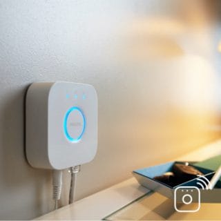 A smart lighting hub with illuminated blue ring is mounted on a wall above a desk, connected by cables, with soft light cast nearby.