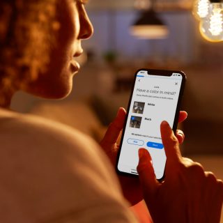 Person using a smartphone to select lighting options in a warmly lit home setting, with pendant lights visible in the background.