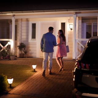 A couple walks toward a well-lit front porch at night, guided by outdoor pathway lights in a residential setting.