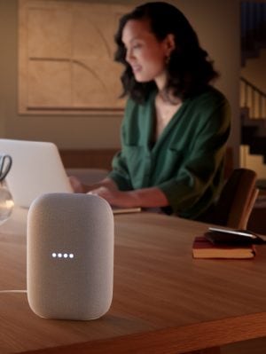 A person working on a laptop at a wooden table with a smart speaker in focus, illuminated by soft indoor lighting.