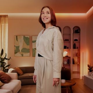 Una mujer de pie en su salón, rodeada de una suave iluminación ambiental blanca con tiras de luces LED y lámparas inteligentes que crean un ambiente relajante.