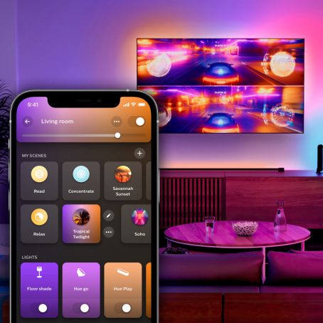 Una sala iluminada con colores vivos y un televisor montado en la pared que muestra un juego de carreras en pantalla dividida. Una superposición de la aplicación Hue muestra la pestaña Inicio.